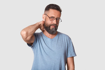 Obraz premium Tired unemotional handsome executive looks firmly at camera, holding his arm behind, making neck massage after hard working day. Middle aged black haired model wearing stylish specs and t shirt.