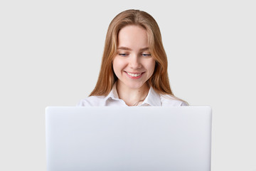 Smiling delighted blue eyed girl sits at table, working on laptop, surfing net, reading funny stories, wathing and reading posts in social networking sites, looks happy, wearing white formal blouse.