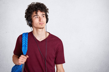 Pensive young guy with headphones on head and blue rucksack, wearing maroon casual t shirt, listens to music, looking thoufhtfully aside, stands isolated over white studio background, Copy space.