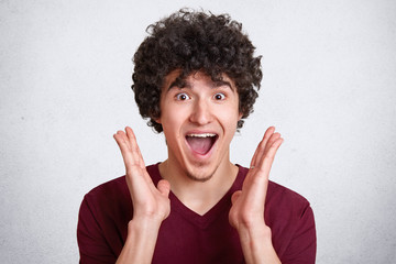 Studio shot of attractive man with dark curly hair, keeps mouth opened, screams something, stands with satisfied facial expression, has high spirit, his dreams come true, poses isolated on white wall.