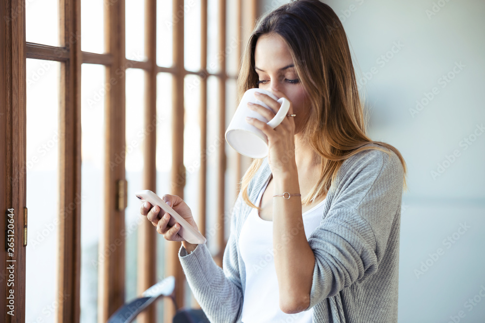 Wall mural Beautiful young woman using her mobile phone while drinking coffee near to the window in the living room at home.