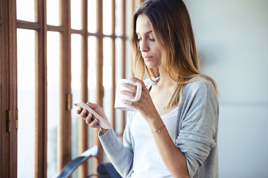 Beautiful young woman using her mobile phone while drinking coffee near to the window in the living room at home.