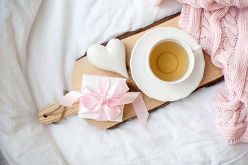 Fototapeta na wymiar White mug with tea, gift box with ribbon on the bed. Breakfast in bed. Cozy. Pink plaid. Cotton.