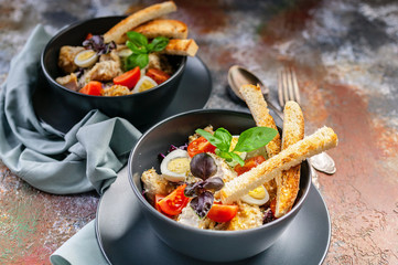 Classic Caesar salad with chicken, quail egg, cherry tomatoes, croutons and basil in dark blue bowls on a rusty background.