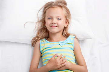 Pretty small child has pleasant look, charming smile, light hair, blue eyes, healthy skin, keeps hands on stomach, dressed in striped pyjamas, looks directly at camera, rests in bedroom alone.
