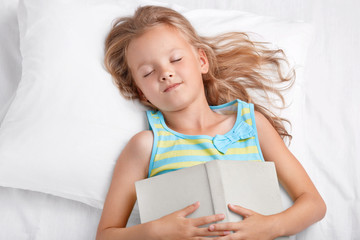View of lovely girl with long light hair sleeps after reading fairy tale, keeps book on stomach, lies in comfortable white bed, has pleasant dreams, enjoys fairytale. Children and relaxation concept