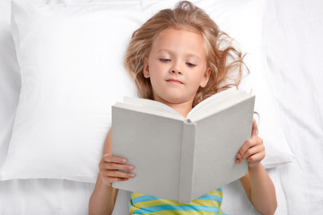 Pretty small preschooler reads attentively interesting story, holds opened book, lies in bed, enjoyes reading before sleep, has good rest. Clever little girl reads fairy tale. Hobby, children concept