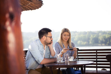 A handsome bearded young man is sitting with his beautiful girl in a restaurant on the shore and he is very bored because she constantly watches something on the phone.