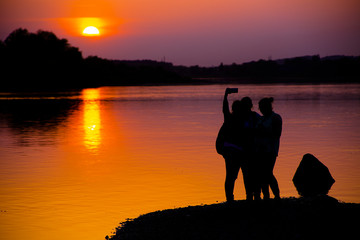 Fototapeta na wymiar Silhouettes of three girls near river at sunset. Silhouettes of young group of people taking a selfie with smartphone.