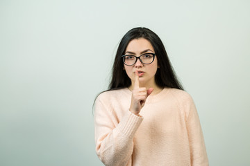 Portrait of a beautiful girl in glasses and a beige sweater in the studio on a white background showing a sign of silence. Concept of secret and mystery.