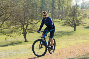 Fototapeta na wymiar Cyclist in shorts and jersey on a modern carbon hardtail bike with an air suspension fork rides off-road on green hills near the forest