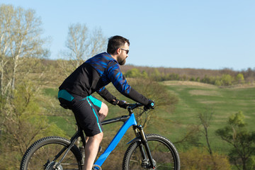 Obraz premium Cyclist in shorts and jersey on a modern carbon hardtail bike with an air suspension fork rides off-road on green hills near the forest
