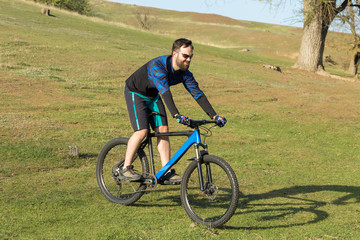 Obraz premium Cyclist in shorts and jersey on a modern carbon hardtail bike with an air suspension fork rides off-road on green hills near the forest