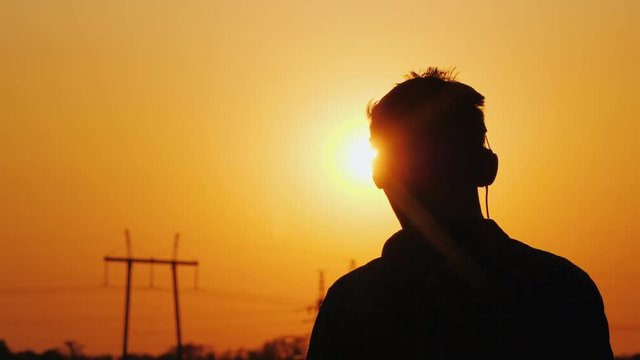 Teenager in headphones listening to music, looking at a beautiful sunset over the city, view from behind