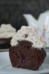 cut chocolate cupcake with a white cheese top stands on a white plate on a black background, behind is a white teapot