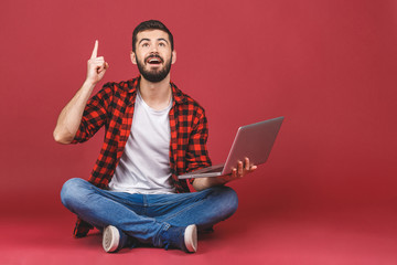 Portrait of a happy young man in white t-shirt holding laptop computer while sitting on a floor and pointing finger up isolated over red background.