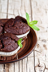 Chocolate dark muffins with mint leaves on brown ceramic plate closeup on rustic wooden table.