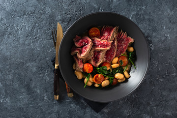 Dinner plate with beef steak with white beans, spinach and tomatoes on dark stone background top view with free co