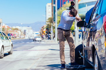 A sweaty tired young entrepreneur is wiping the sweat off his forehead while trying to change a flat tire on a beautiful sunny day