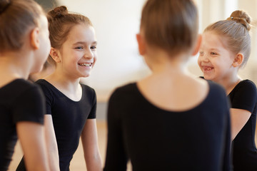 Group of cute girls giggling while sitting in circle during ballet class in studio lit by warm...