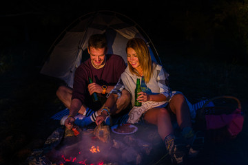 Fototapeta premium Young couple baking sausages on the campfire and drinking beer in the forest hill in the dusk