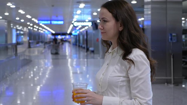 Woman drink fresh orange juice, half length shot in empty airport hall, blurred background. Lady hold plastic glass and make gulp of freshly squeezed drink from vending machine.