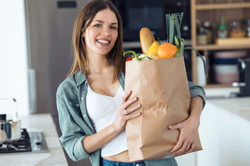 Beautiful young woman looking at camera while holding shopping bag with fresh vegetables in the kitchen at home.