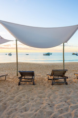 Beach with wooden chairs, sun loungers and tent with boats and sunset in the background.Concept of vacations, peace and relaxation. Ponta do Corumbau, Bahia, Brazil.