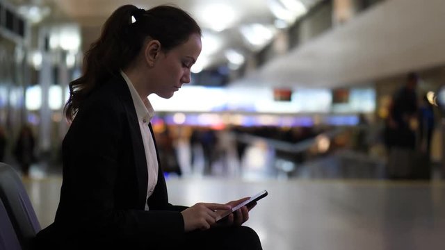 Young adult woman browse through images on smartphone, dim airport hall area, blurred background. Lady spend time waiting for flight boarding, behold to mobile device