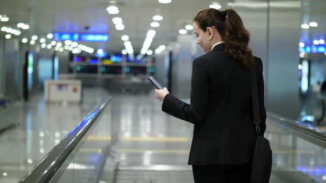 Lady read information using mobile device, stay on moving travellator, going to boarding gate. Blurred airport hall on background, dim lighting at evening time