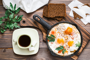 Ready-to-eat breakfast: shakshuka from fried eggs with tomatoes and parsley in a pan, bread with butter and coffeee on a wooden table. Top view