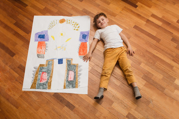 the child with the big drawing of the robot playing  lies on a floor