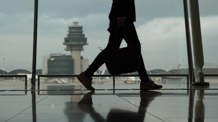 Business woman with handbag and smartphone walk at terminal against window, silhouetted shot of legs, slow motion. Modern HKG airport, tower and apron seen outdoors, cloudy sky