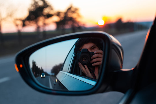 Young photographer taking selfie picture in a mirror of car with professional dslr camera, on background of sunset.