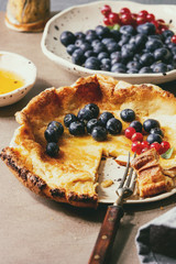 Started eaten fresh baked Dutch baby pancake in ceramic plate with blackberry and red currant berries, bowl of honey, jug of cream, vintage cutlery over beige kitchen table. Close up
