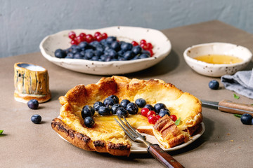 Started eaten fresh baked Dutch baby pancake in ceramic plate with blackberry and red currant berries, bowl of honey, jug of cream, vintage cutlery over beige kitchen table.