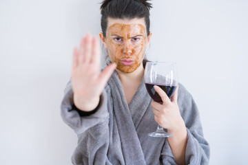 Beautiful woman wearing cosmetic facial mask as skincare treatment drinking glass of wine with open hand doing stop sign with serious and confident expression, defense gesture