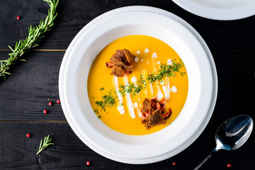 Pumpkin and carrot soup with cream and seasoning in white bowl on black wooden background, top view