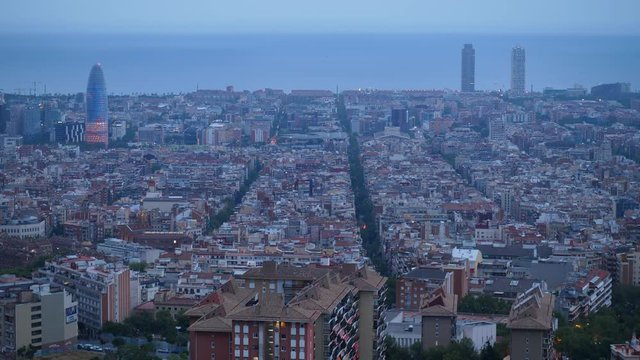 Aerial perspective of Barcelona at dusk, straight streets crossing dense build area, Torre Glories seen on left, quiet sea waters on background.