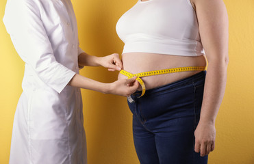 Doctor measuring waist of overweight woman on color background, closeup