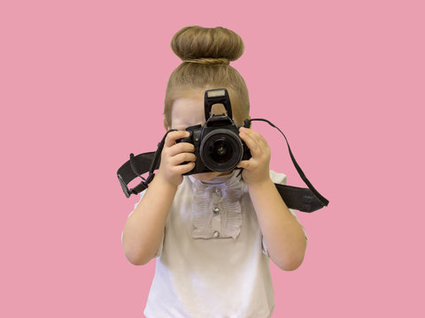 A little girl in business clothes holds a reflex camera in her hands in front of her face. The child wants to take a picture. The baby holds her finger on the shutter button.