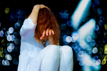 A white dressed woman is sitting in a dark room. Her hair covers her face. She suffers from sensory overload. Concept of health and illness.