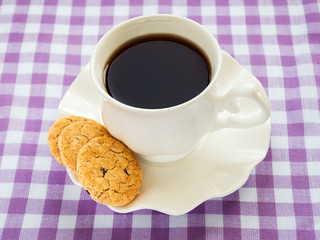 White porcelain saucer with wavy edge and cup of tea and sweet cookies on a checkered white purple tablecloth. Oat cookies with pieces of dark chocolate.