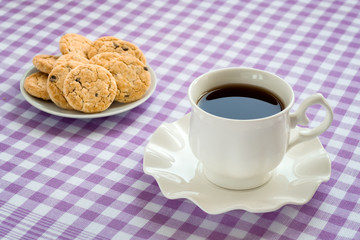 Porcelain cup with tea on saucer with wavy edge and oat cookies with pieces of dark chocolate on a checkered white purple tablecloth.