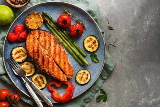 Grilled salmon fish with vegetables zucchini, asparagus, tomato, sweet pepper on a plate, gray background. Top view, flat lay,copy space