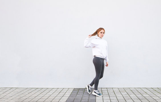 Portrait of a sports girl in white sportswear and leggings, posing on the background of a white wall. Fitness girl is training on a white background, looking into the camera. Sports concept.