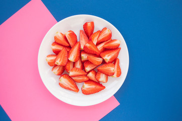 Fresh juicy cut strawberries on a white plate on blue and pink background with copyspace. Flat lay style.