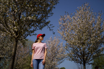 Successful business woman enjoys her leisure free time in a park with blossoming sakura cherry trees wearing jeans, pink t-shirt and a fashion red cap