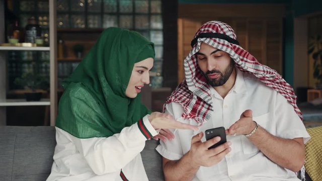 Emirati young couple use phone talk smile sitting on sofa at home conversation atrium female lifestyle culture arab indoors beard region smartphone downtown emirates tourism close up slow motion
