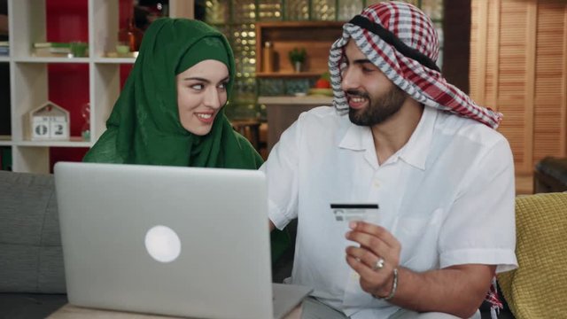 Attractive emirati young couple use laptop credit card shop talk smile sitting on sofa at home conversation atrium female lifestyle culture arab indoors beard region emirates close up slow motion